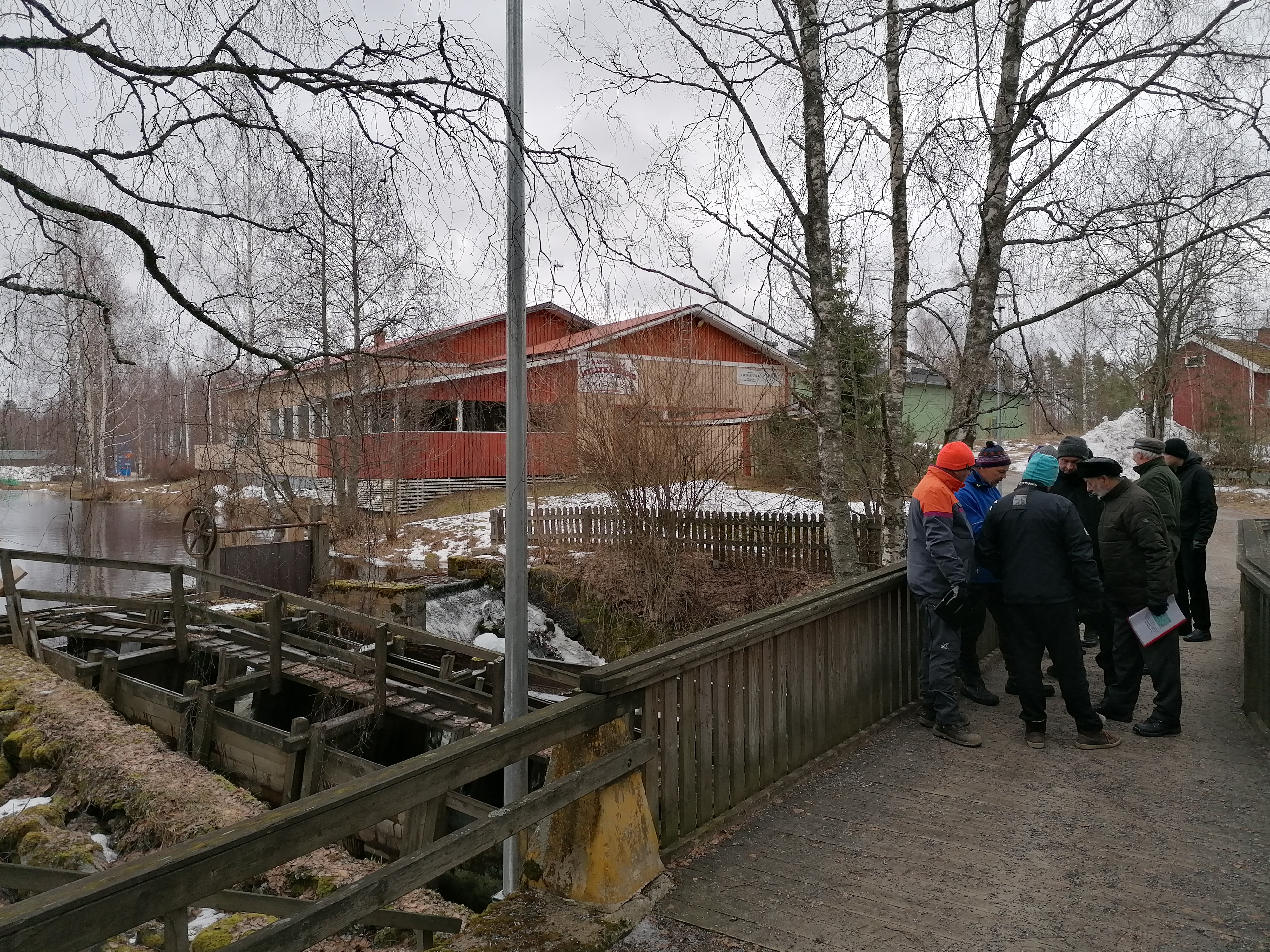 Näkymä Myllysillalle, jossa padon purkua valmistelevan hankkeen osallisia on kokoontunut työmaakatselmukseen. Vasemmalla vanha Myllypato ja puinen kalatie, taustalla häämöttää patoallas. Talvikuva.