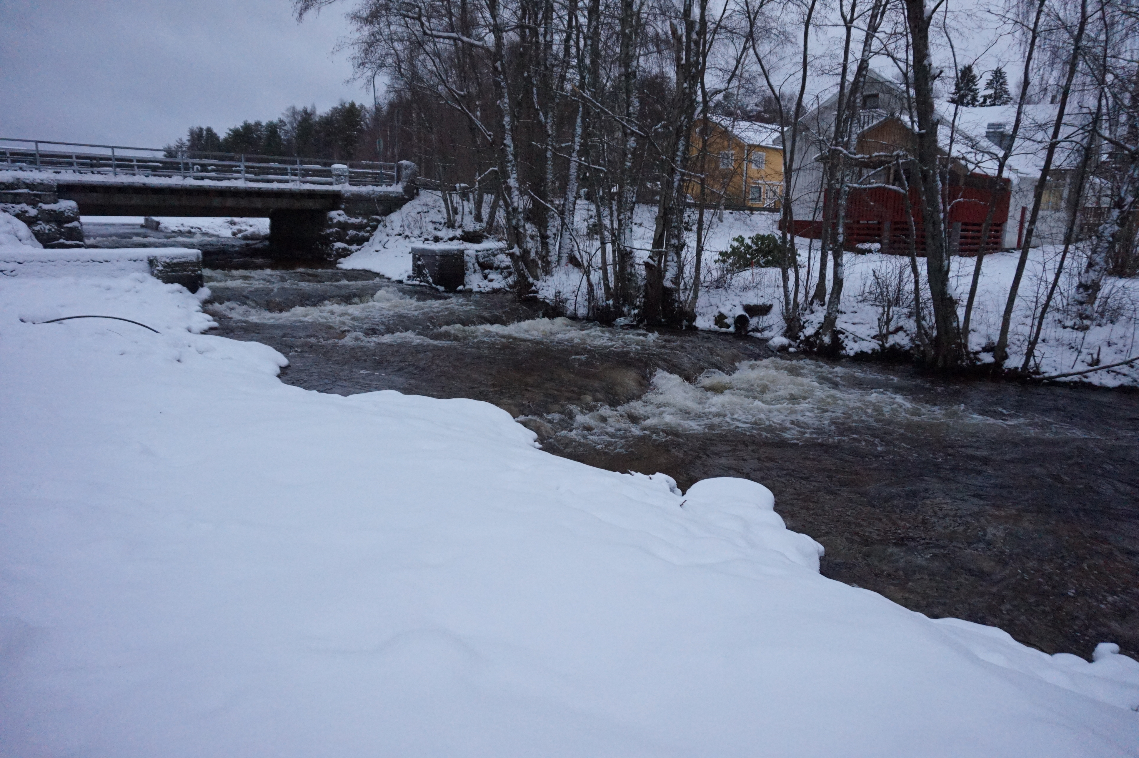 ETualalla lunta, sitten tummana virtaava koski ja taustalla rannalla puita ja taustalla taloja.
